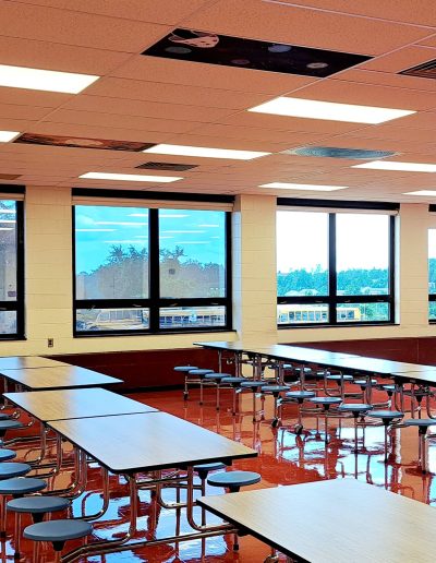 Interior of school cafeteria with several tables and chairs lined up in rows with rectangular windows on wall