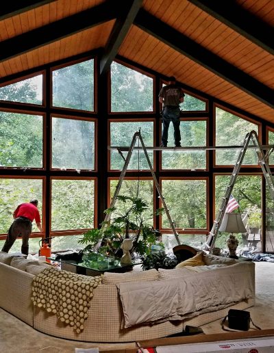 Window washers cleaning large wall covered in square windows using ladders inside modern home with vaulted wooden ceilings