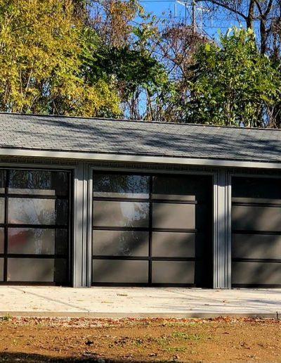 Grey outdoor shed with three large rectangular windows divided into panels in front of trees
