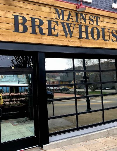 Outside front view of Main St Brewhouse location with wooden sign, grey painted brick, mirrored glass wall divided with black metal and glass door