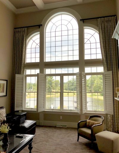 Large white divided windows with white shutters and large curved divided windows above in formal living room overlooking pond and trees