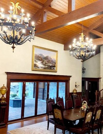 Interior view of formal dining room with two chandeliers hanging from wooden rafters above ornate wooden table and chairs showcasing large glass doors
